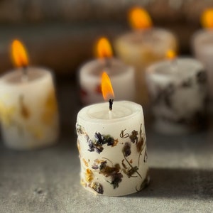 K&ouml;nnte beinhalten: Ein Satz kleiner wei&szlig;er Kerzen mit getrockneten Blumen, die in das Wachs eingebettet sind. Die Kerzen sind angez&uuml;ndet und die Flammen sind sichtbar.