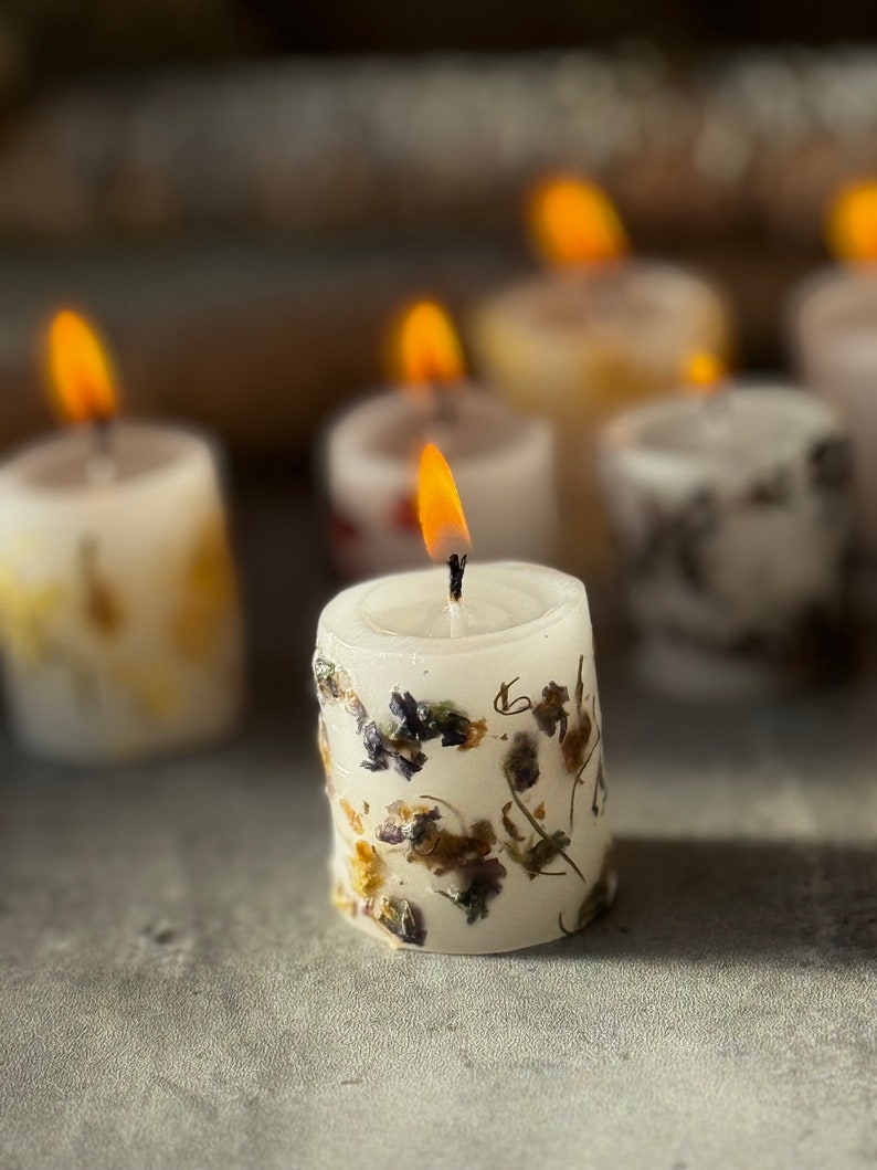 K&ouml;nnte beinhalten: Ein Satz kleiner wei&szlig;er Kerzen mit getrockneten Blumen, die in das Wachs eingebettet sind. Die Kerzen sind angez&uuml;ndet und die Flammen sind sichtbar.