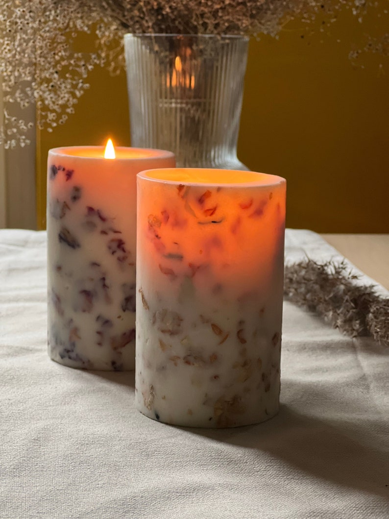 May include: Two white pillar candles with embedded dried flowers and a warm glow. The candles are on a white linen tablecloth.