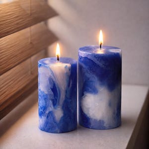 May include: Two lit, cylindrical candles with a marbled blue and white design. The candles are placed side-by-side on a white surface, with a wooden blind in the background. The flames are bright yellow.
