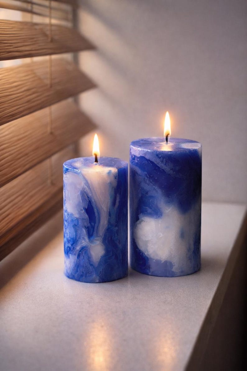 May include: Two lit, cylindrical candles with a marbled blue and white design. The candles are placed side-by-side on a white surface, with a wooden blind in the background. The flames are bright yellow.