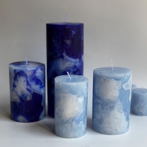 May include: A collection of pillar candles in varying sizes, featuring a marbled design in shades of blue and white. The candles have a cylindrical shape and are arranged on a white surface. The tallest candle is approximately 6 inches tall.