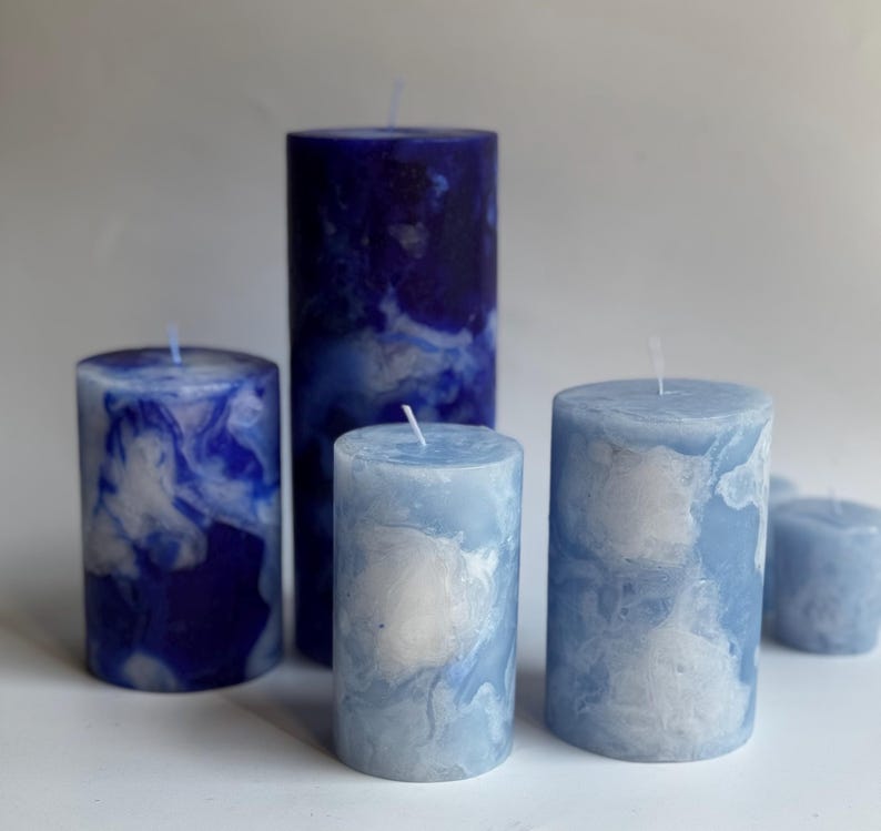 May include: A collection of pillar candles in varying sizes, featuring a marbled design in shades of blue and white. The candles have a cylindrical shape and are arranged on a white surface. The tallest candle is approximately 6 inches tall.