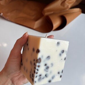 K&ouml;nnte beinhalten: Eine quadratische, cremefarbene Kerze mit eingebetteten dunkelbraunen Kaffeebohnen. Die Kerze hat einen einzigen Docht. Der Hintergrund umfasst braunes Papier und eine wei&szlig;e Oberfl&auml;che. Die Kerze wird von einer Hand gehalten.