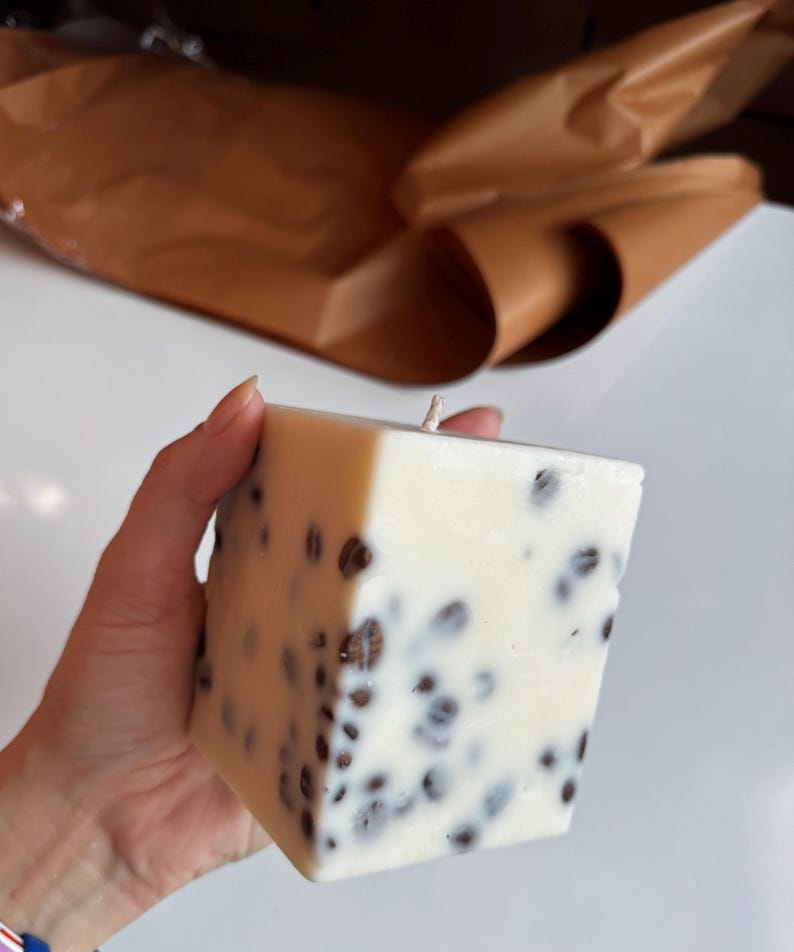 K&ouml;nnte beinhalten: Eine quadratische, cremefarbene Kerze mit eingebetteten dunkelbraunen Kaffeebohnen. Die Kerze hat einen einzigen Docht. Der Hintergrund umfasst braunes Papier und eine wei&szlig;e Oberfl&auml;che. Die Kerze wird von einer Hand gehalten.