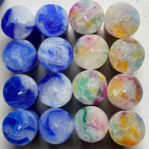 May include: A collection of sixteen round candles. The candles are arranged in a grid pattern and feature a variety of colors, including blue, green, pink, and yellow. Each candle has a small wick in the center.