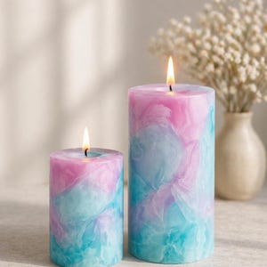 Könnte beinhalten: Zwei brennende Säulenkerzen mit einem marmorierten Design in Rosa-, Blau- und Lilatönen. Die Kerzen sind zylindrisch und haben ein weiches, leuchtendes Aussehen. Im Hintergrund steht eine kleine Vase mit weißen Blumen.