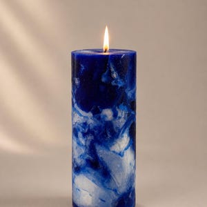 May include: A lit, cylindrical pillar candle with a marbled design in shades of blue and white. The candle has a smooth surface and a small, flickering flame at the top. The candle is a decorative item.