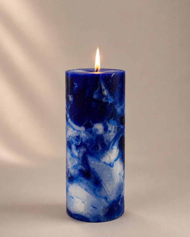 May include: A lit, cylindrical pillar candle with a marbled design in shades of blue and white. The candle has a smooth surface and a small, flickering flame at the top. The candle is a decorative item.
