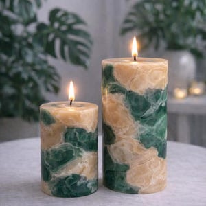 Op de afbeelding: Twee brandende pilaar kaarsen met een marmeren ontwerp. De kaarsen hebben een mix van groene en beige tinten, waardoor een natuurlijk, steenachtig uiterlijk ontstaat. De kaarsen zijn cilindrisch en staan op een lichtgekleurd oppervlak, met groen op de achtergrond.