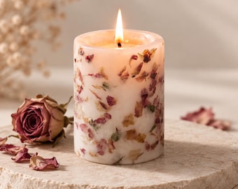 May include: A cylindrical white wax candle with a lit wick, featuring embedded dried pink and red rose petals throughout its exterior. A single dried rosebud and scattered loose petals are positioned near the base of the candle.