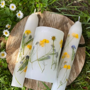 Handmade Unity candles rest on a thick, round, wooden base. Candles are white, made from paraffin wax and have real wild flowers pressed into them. Flowers looks like they are painted on the candles. Everything is lying down on a meadow.