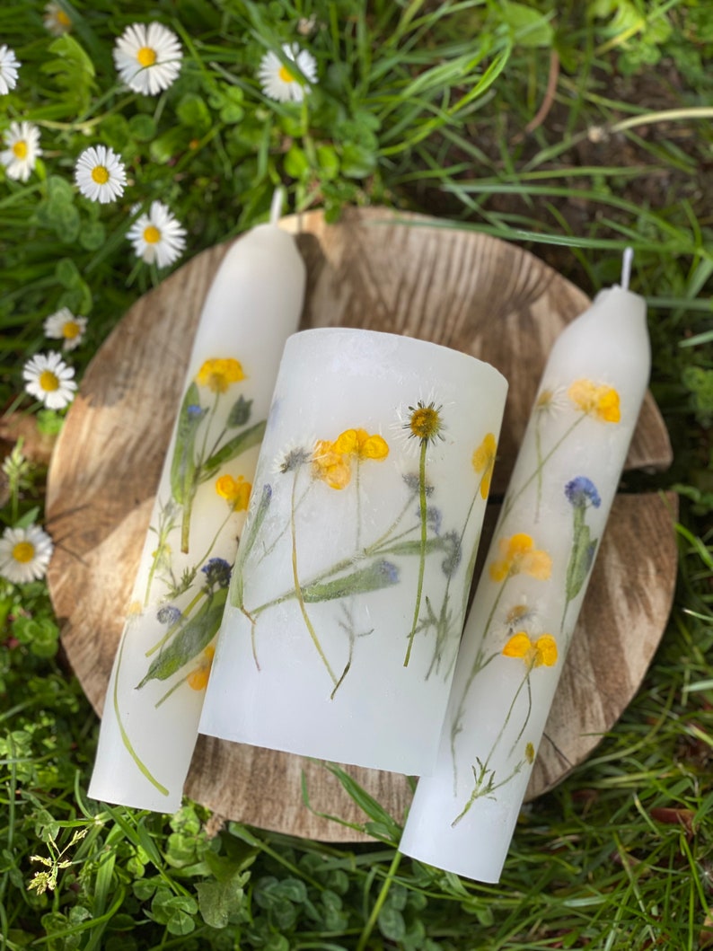 Handmade Unity candles rest on a thick, round, wooden base. Candles are white, made from paraffin wax and have real wild flowers pressed into them. Flowers looks like they are painted on the candles. Everything is lying down on a meadow.