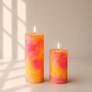 May include: Two cylindrical pillar candles with a marbled pattern in vibrant shades of pink, orange, and yellow. Both candles are lit, featuring a single burning wick at the centre of each flat top, creating a warm glow against the wax surface.