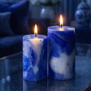 May include: Two lit pillar candles with a marbled blue and white design. The candles are cylindrical and have a wax-like appearance. The flames are bright yellow-orange. The background is a dark blue, creating a calming ambiance.