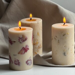 May include: Three candles with dried flower petals embedded in the wax. The candles are white and have a natural, rustic look. The candles are lit and the flames are visible.