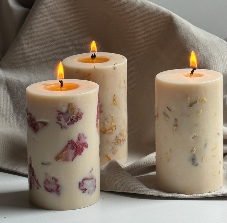 May include: Three candles with dried flower petals embedded in the wax. The candles are white and have a natural, rustic look. The candles are lit and the flames are visible.