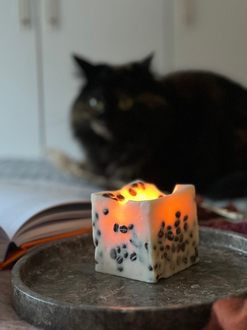 K&ouml;nnte beinhalten: Eine quadratische, wei&szlig;e Kerze mit eingebetteten Kaffeebohnen, die auf einem grauen Marmortablett steht. Die Kerze ist angez&uuml;ndet und strahlt ein warmes Licht aus. Eine schwarze Katze ist im Hintergrund verschwommen, zusammen mit einem offenen Buch.