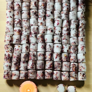 K&ouml;nnte beinhalten: Eine gro&szlig;e Sammlung wei&szlig;er Teelichter mit getrockneten Rosenbl&uuml;ten, die in das Wachs eingebettet sind. Die Kerzen sind in einem Gittermuster auf einer Holzoberfl&auml;che angeordnet. Im Vordergrund befinden sich drei weitere Kerzen, von denen eine brennt.