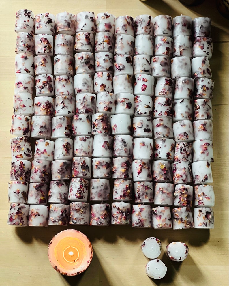K&ouml;nnte beinhalten: Eine gro&szlig;e Sammlung wei&szlig;er Teelichter mit getrockneten Rosenbl&uuml;ten, die in das Wachs eingebettet sind. Die Kerzen sind in einem Gittermuster auf einer Holzoberfl&auml;che angeordnet. Im Vordergrund befinden sich drei weitere Kerzen, von denen eine brennt.