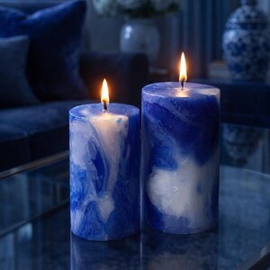 May include: Two lit pillar candles with a marbled blue and white design. The candles are cylindrical and sit on a reflective surface. The flames are bright yellow against the dark blue background.