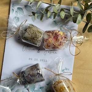 K&ouml;nnte beinhalten: Vier kleine, klare Glaskerzenbeh&auml;lter mit verschiedenen Trockenblumengebinden darin. Die Beh&auml;lter sind in durchsichtige Cellophanfolie verpackt und mit Schnur gebunden. Der Text "Olivia & Johan 09.25.2022" ist auf einer wei&szlig;en Karte hinter den Beh&auml;ltern gedruckt.