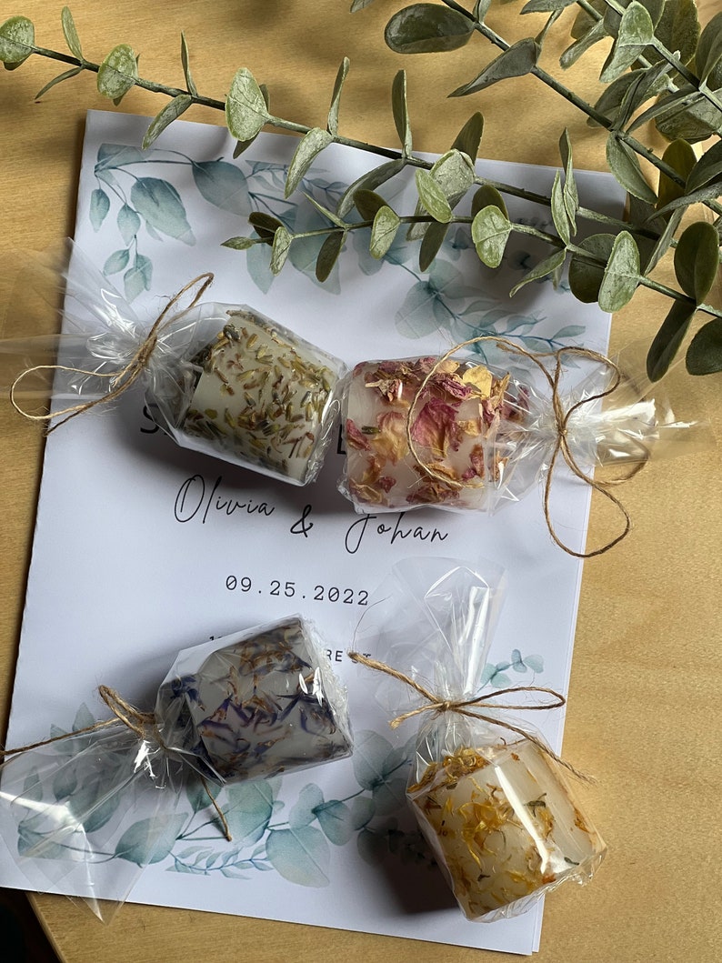 K&ouml;nnte beinhalten: Vier kleine, klare Glaskerzenbeh&auml;lter mit verschiedenen Trockenblumengebinden darin. Die Beh&auml;lter sind in durchsichtige Cellophanfolie verpackt und mit Schnur gebunden. Der Text "Olivia & Johan 09.25.2022" ist auf einer wei&szlig;en Karte hinter den Beh&auml;ltern gedruckt.