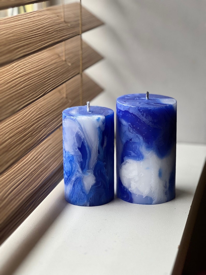 May include: Two pillar candles with a marbled design in shades of blue and white. The candles are cylindrical, with a wick at the top. The background includes wooden blinds.