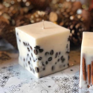 K&ouml;nnte beinhalten: Eine quadratische, cremefarbene Kerze mit eingebetteten Kaffeebohnen. Die Kerze hat eine glatte, wachsartige Textur und einen einzelnen Docht. Die Kaffeebohnen sind dunkelbraun und verleihen der Kerze ein dekoratives Element. Ein Einrichtungsgegenstand.