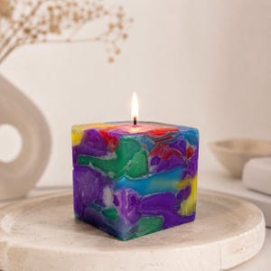 Pode incluir: Uma vela quadrada com um design abstrato colorido. A vela apresenta uma mistura de tons roxos, azuis, verdes, vermelhos e amarelos. Um pavio aceso fica em cima da vela, que repousa sobre uma bandeja redonda branca.