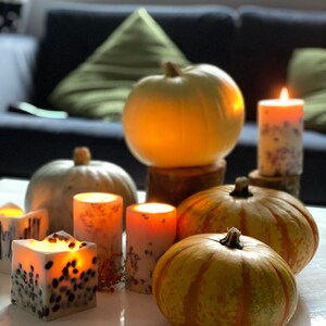 K&ouml;nnte beinhalten: Eine herbstliche Dekoration mit K&uuml;rbissen und brennenden Kerzen. Die K&uuml;rbisse sind in verschiedenen Gr&ouml;&szlig;en und Farben, einige wei&szlig; und andere orange mit gr&uuml;nen Streifen. Die Kerzen sind zylindrisch und quadratisch, mit floralen und gesprenkelten Mustern.