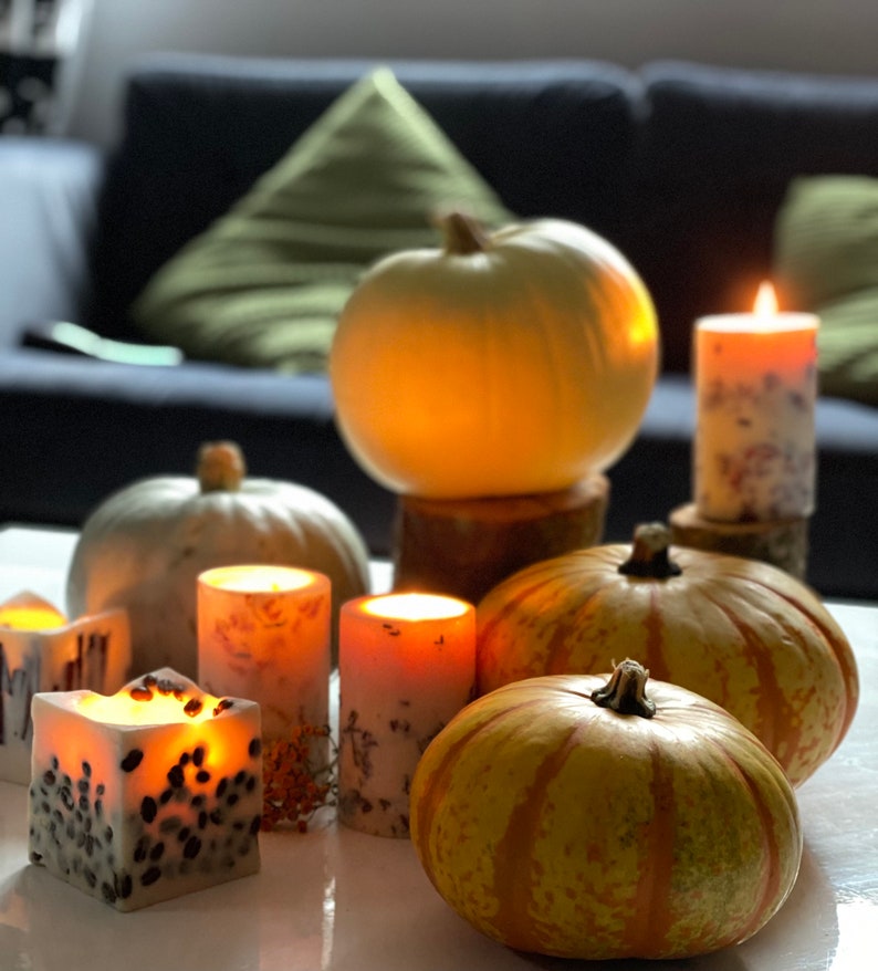 K&ouml;nnte beinhalten: Eine herbstliche Dekoration mit K&uuml;rbissen und brennenden Kerzen. Die K&uuml;rbisse sind in verschiedenen Gr&ouml;&szlig;en und Farben, einige wei&szlig; und andere orange mit gr&uuml;nen Streifen. Die Kerzen sind zylindrisch und quadratisch, mit floralen und gesprenkelten Mustern.