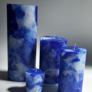 May include: Four blue and white marbled pillar candles of varying heights. The candles have a marbled effect with shades of blue and white. Each candle has a single wick at the top, and they are arranged on a white surface.