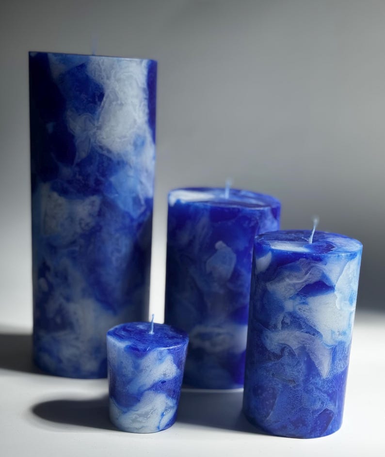 May include: Four blue and white marbled pillar candles of varying heights. The candles have a marbled effect with shades of blue and white. Each candle has a single wick at the top, and they are arranged on a white surface.