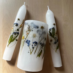 White Unity candles with real flowers pressed into them. It looks like flowers are painted on the candles. Two thin long candles are lying on the sides and lower thick candle is placed in the middle of them. Candles rest on a wooden, oak table.