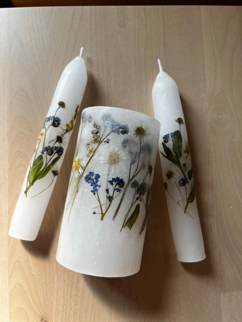 White Unity candles with real flowers pressed into them. It looks like flowers are painted on the candles. Two thin long candles are lying on the sides and lower thick candle is placed in the middle of them. Candles rest on a wooden, oak table.