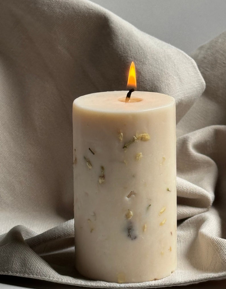 May include: A white candle with dried flowers embedded in the wax. The candle is lit and the flame is visible. The candle is sitting on a beige fabric surface.