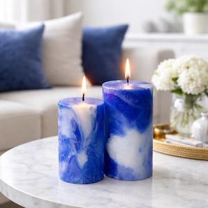 May include: Two lit blue and white marbled pillar candles of different sizes sit on a marble table. The candles have a swirling pattern of blue and white, with a soft glow from the flames. The background includes a white sofa and blue pillows.
