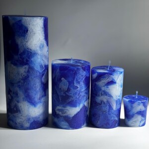 May include: Four blue pillar candles of varying heights, with a marbled white and blue design. The candles are cylindrical, with a wick at the top. The tallest candle is on the left, with the smallest on the right.