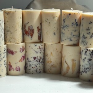 May include: A collection of 14 cylindrical candles with a cream-colored base and embedded dried flowers. The flowers are in shades of purple, pink, and brown.