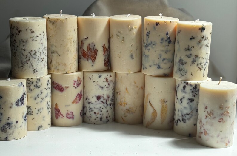 May include: A collection of 14 cylindrical candles with a cream-colored base and embedded dried flowers. The flowers are in shades of purple, pink, and brown.