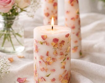 May include: A cylindrical pillar candle and two tapered candles made of white wax, each embedded with dried pink and yellow flower petals. The pillar candle is lit, and the two tapered candles are also lit, standing upright with visible flames.