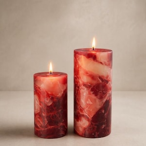 May include: Two pillar candles with a marbled red and cream design. The candles are lit, with flames at the top. The larger candle is taller than the smaller one. The candles are on a neutral background.