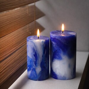 May include: Two lit, cylindrical candles with a marbled blue and white design. The candles are placed on a white surface near a window with wooden blinds. The flames are yellow.