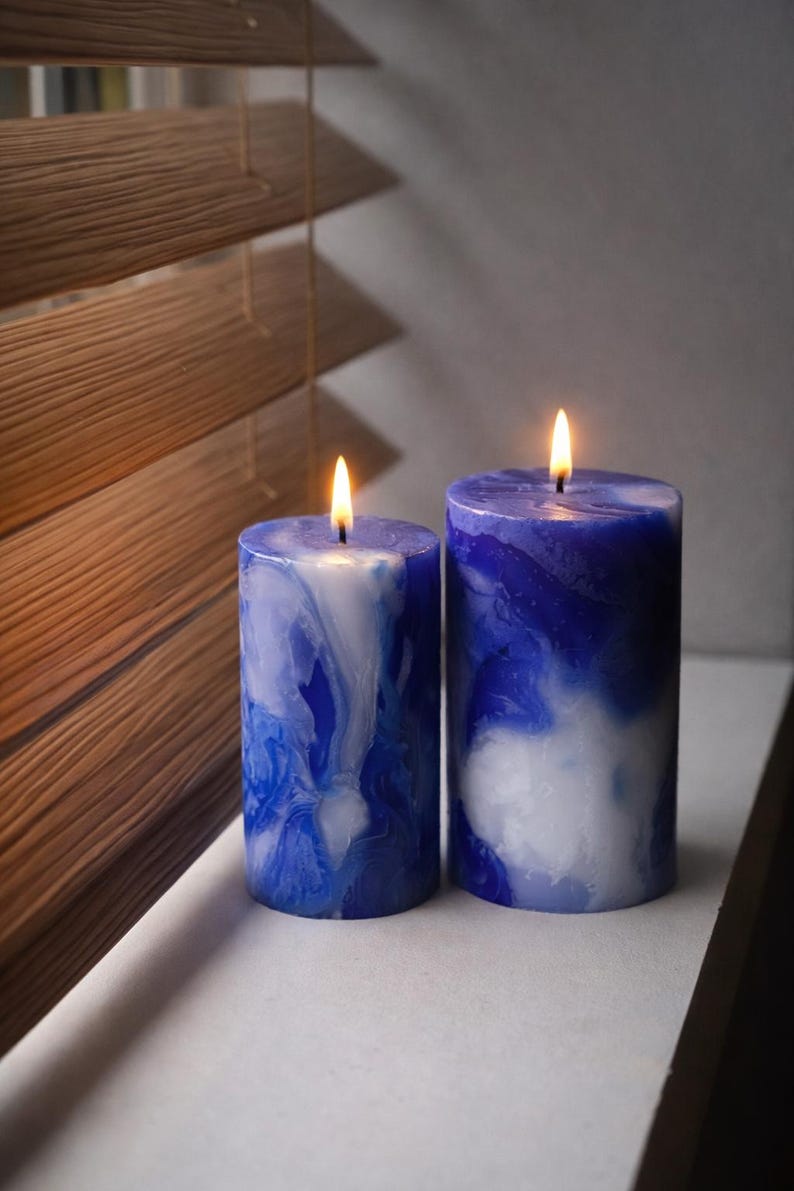 May include: Two lit, cylindrical candles with a marbled blue and white design. The candles are placed on a white surface near a window with wooden blinds. The flames are yellow.