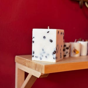 K&ouml;nnte beinhalten: Eine quadratische, wei&szlig;e Kerze mit dunklen Kaffeebohnen. Die Kerze steht auf einem Holzregal vor einer roten Wand. Weitere kleinere Kerzen sind im Hintergrund zu sehen.