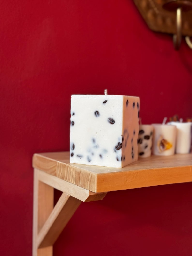K&ouml;nnte beinhalten: Eine quadratische, wei&szlig;e Kerze mit dunklen Kaffeebohnen. Die Kerze steht auf einem Holzregal vor einer roten Wand. Weitere kleinere Kerzen sind im Hintergrund zu sehen.