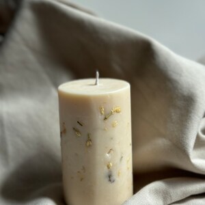 May include: A white pillar candle with dried flowers and gold flakes embedded in the wax. The candle is sitting on a light brown fabric surface.
