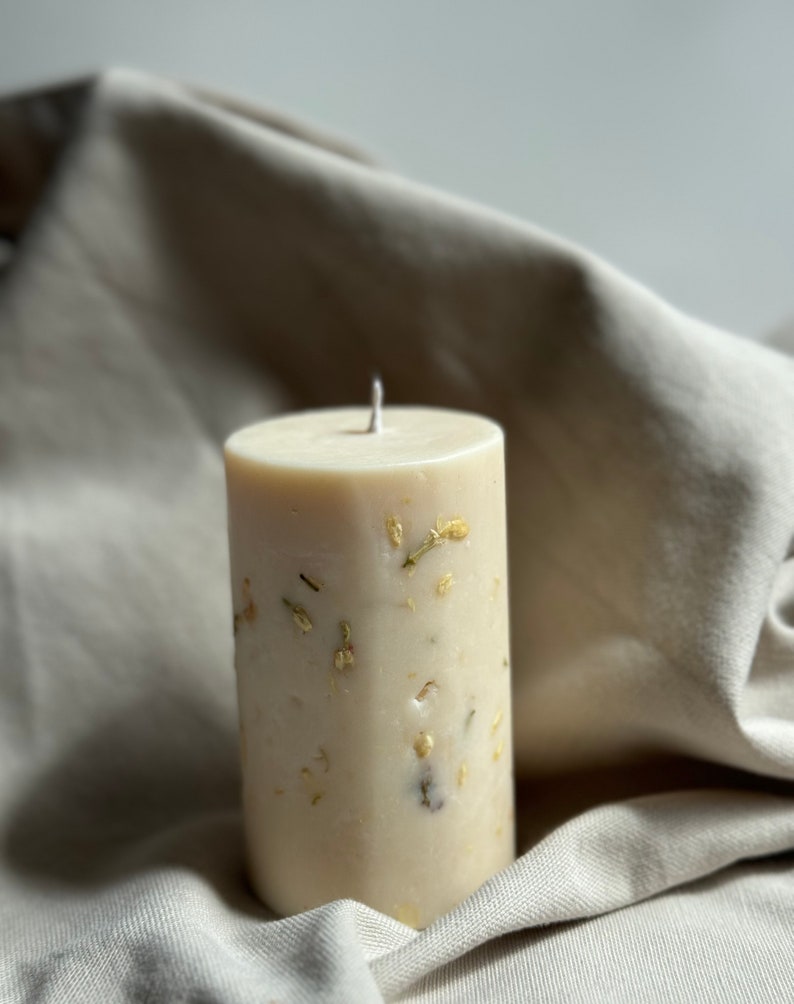 May include: A white pillar candle with dried flowers and gold flakes embedded in the wax. The candle is sitting on a light brown fabric surface.