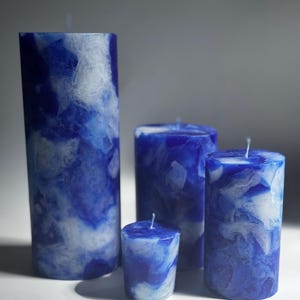 May include: Four blue and white marbled pillar candles of varying heights. The candles have a marbled effect with shades of blue and white. The wicks are visible on top. The candles are cylindrical and stand on a white surface.